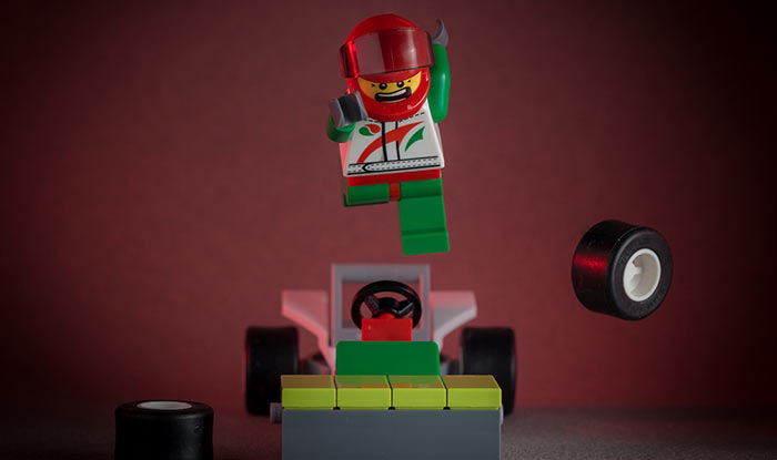 A plastic LEGO man in race car driver attire flies through the air over his race car that has lost two wheels.